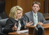 Cheryl Hennen sits at the testifiers' table and speaks to someone out of view while gesturing with her hands. Representative Lippert sits next to her and watches her as she speaks.